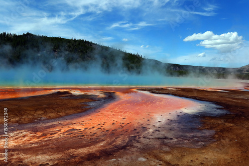 A very beautiful landscape of a large hot lake from which a blue haze rises. There are several small clouds over the lake. In the foreground is bright red mud from the source.