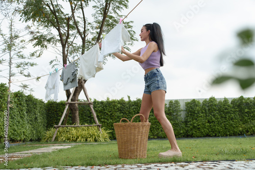 Asian female doing laundry outdoors on a sunny day, placing clothes on the line in her home garden.