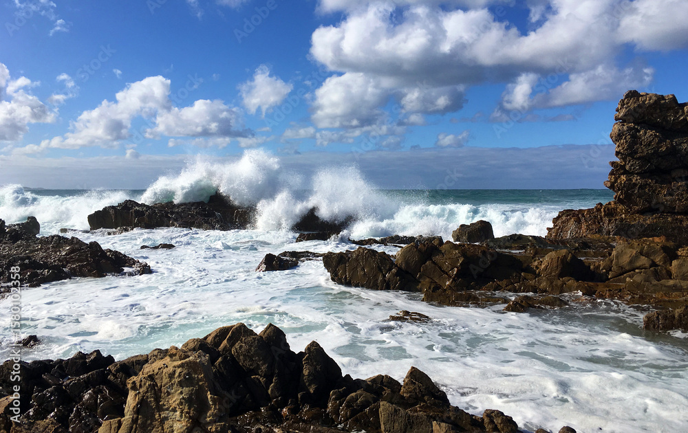 Obraz premium Wild seas at Mallacoota, Australia. Waves crash over rocks at Bastion Point, churning the water into a mass of white foam that swirls around the rocky headland under a vivid blue sky.