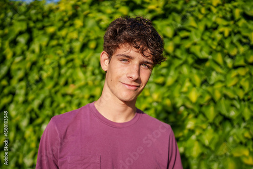 Young student with brown curly hair wearing purple t shirt, smiling near green ivy backdrop