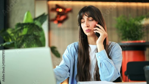 Professional young businesswoman wearing a light blue shirt, conversing on her smartphone while using a laptop in a modern office. She looks focused and cheerful.