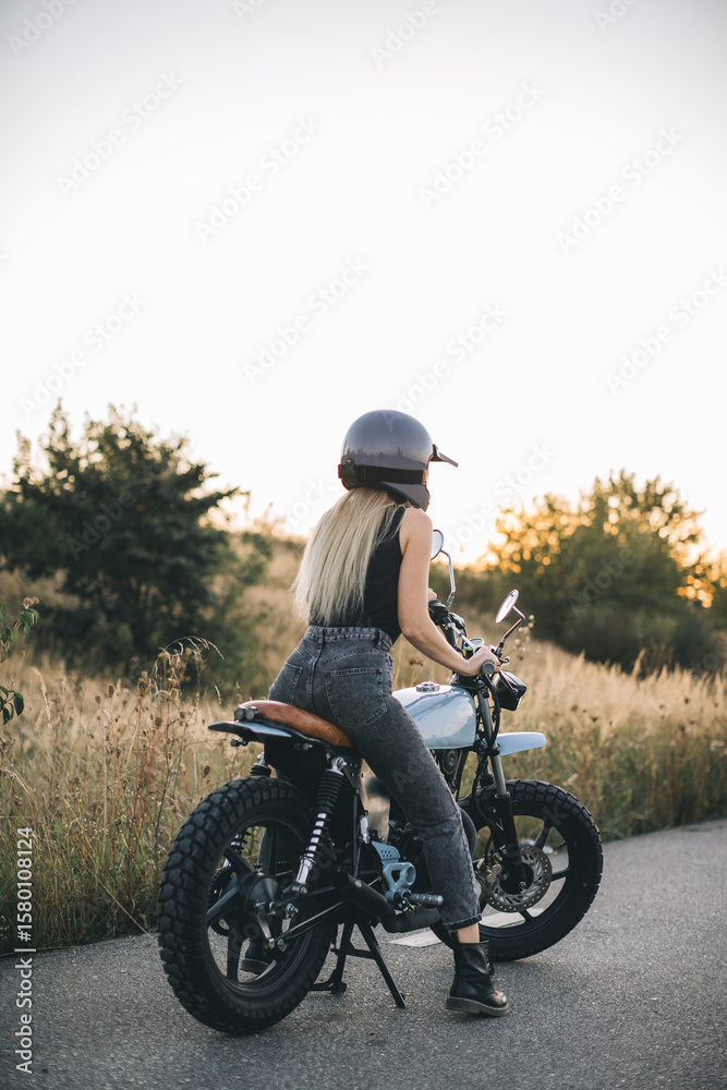 Fototapeta premium Young blonde woman riding custom motorcycle at sunset