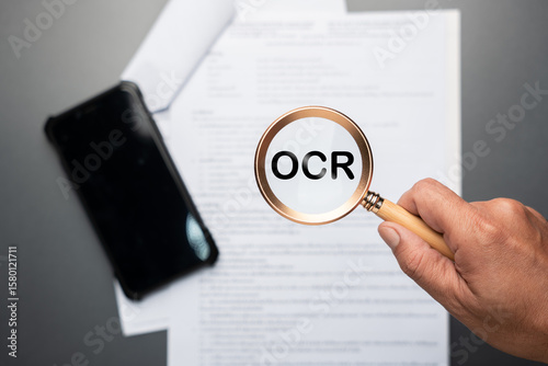 Hand holding magnifying glass with OCR text over document paper and smartphone. Symbolizes optical character recognition, text extraction, digitizing printed documents, and digital data conversion