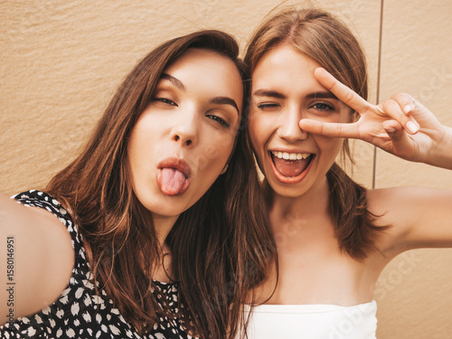Two young smiling hipster women in summer clothes. Female taking selfie self portrait photos on smartphone.Models posing on street background near yellow wall.Female showing positive face emotions