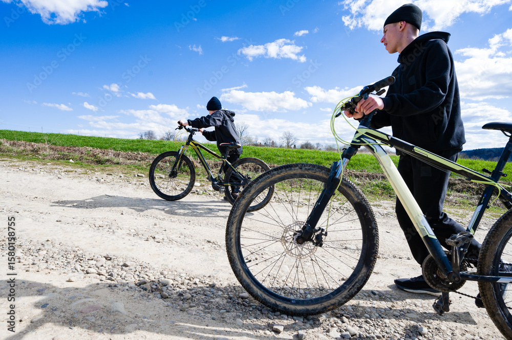 Obraz premium Two cyclists enjoying a sunny day on a dirt road. One cycles, the other walks alongside.