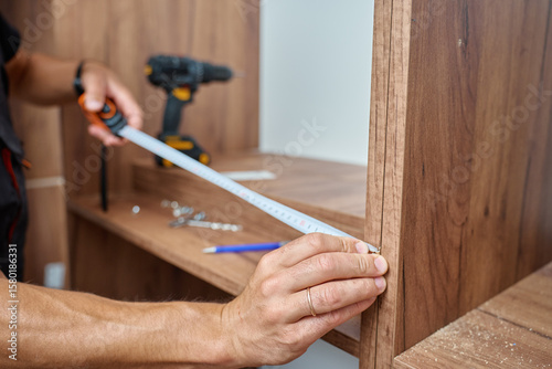 Close-up of man measuring wooden wardrobe with tape measure during furniture assembly, with tools and screws on shelf in background. Concept of home renovation and DIY furniture project