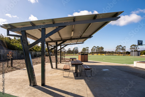 Modern outdoor shelter structure with picnic tables beside a public basketball court in suburban Australia. The covered seating provides shade. Community infrastructure and recreational design