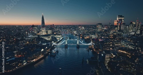 London: Aerial view of city illuminated skyline at sunset, iconic Tower Bridge, The Shard, and night cityscape stretching along the River Thames. Drone flight footage zoom out panorama. Blue toning