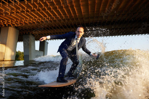 Young man surfer in classic suit rides on the wave. Happy clerk escaped from stuffy office to take up active sport. Best summer leisure after routine work. Film grain effect.