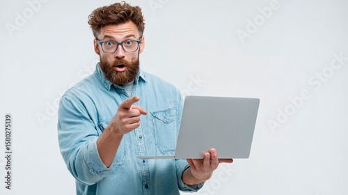 Man with beard in glasses holds laptop pointing with surprised expression denim shirt