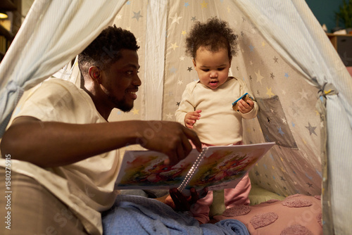 Fotografía Black young adult man spending paternity leave reading storybook to baby inside