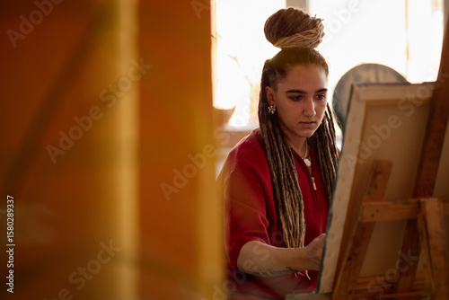 Εκτύπωση καμβά Young adult Caucasian woman with long dreadlocks painting on canvas, sitting at