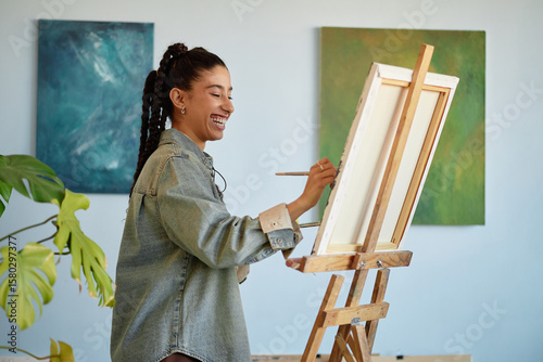 Young adult woman smiling while painting on canvas at easel, holding paintbrush in hand, standing in front of abstract artwork, engaging in creative activity