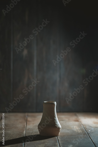 Handcrafted ceramic vase rests on a rustic wooden surface, illuminated by soft, dramatic lighting. The dark background enhances the focus on the vase's unique shape and texture