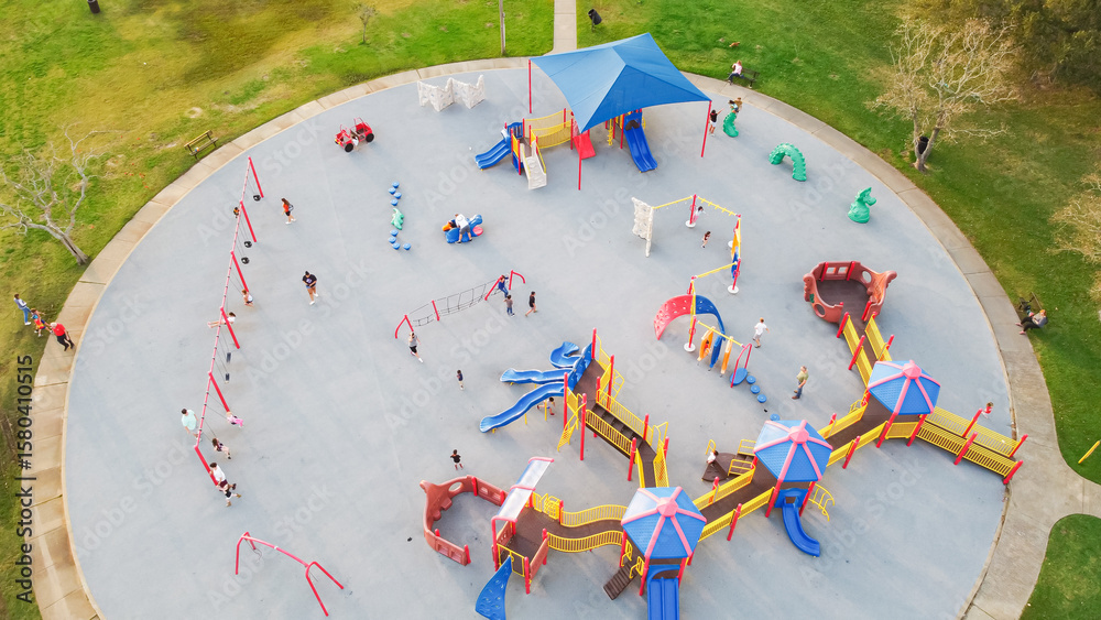 Fototapeta premium Kids swing, climb, and slide on a rounded concrete playground pad while parents supervise. Bright equipment and green lawns surround this lively outdoor scene in Clear Lake Park, Seabrook, Texas