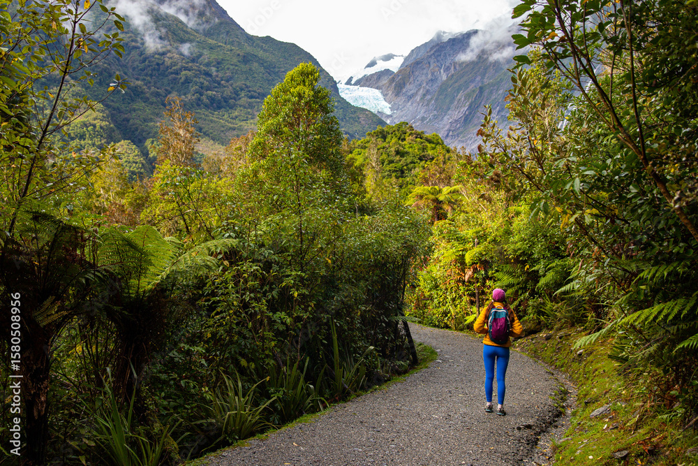 Fototapeta premium girl in yellow jacket walking through dense rainforest with view of Franz Josef Glacier, Westland Tai Poutini National Park, West Coast, South Island, New Zealand