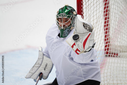 Hockey goalie in full gear makes a dramatic glove save as puck flies mid-air near goal on icy rink. photo