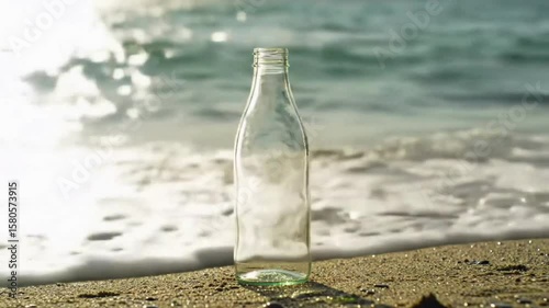 A clear glass bottle stands upright on a sandy beach with ocean waves in the background sunlight