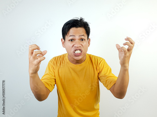 Fotografie A young Asian man wearing a yellow shirt displays strong frustration and anger with vivid expressions, standing against a white background, conveying intense emotions