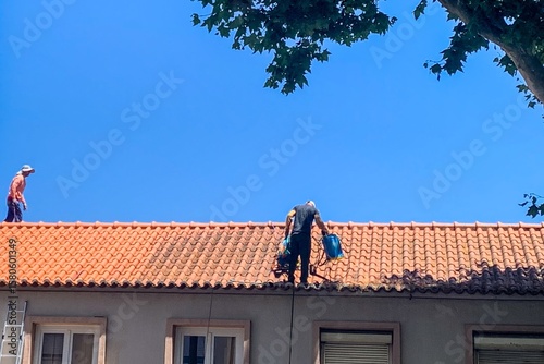 Two diligent workers, one Hispanic and one European, scale terracotta rooftops under azure skies, echoing International Workers' Day valor