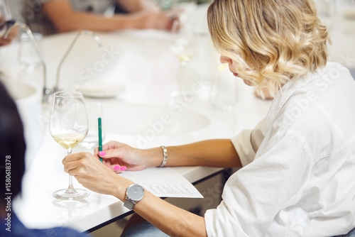 Young handsome looking woman tasting white wine and writing notes to degustation card. Professional sommelier studies wine on lecture about varieties and geographical wine. 