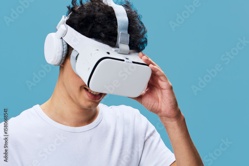young man wearing virtual reality headset and wireless headphones enjoying immersive technology against pastel blue background, smiling and engaged in digital experience