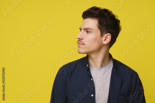 A young man with dark hair and a mustache looks thoughtfully to the side, standing against a bright yellow backdrop. He is wearing a navy blue shirt over a gray t-shirt