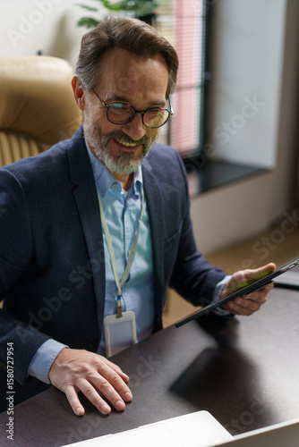 A cheerful man with a beard is enjoying a virtual meeting at his stylish office. Dressed in a smart suit, he holds a tablet and smiles, immersed in the conversation as sunlight filters through
