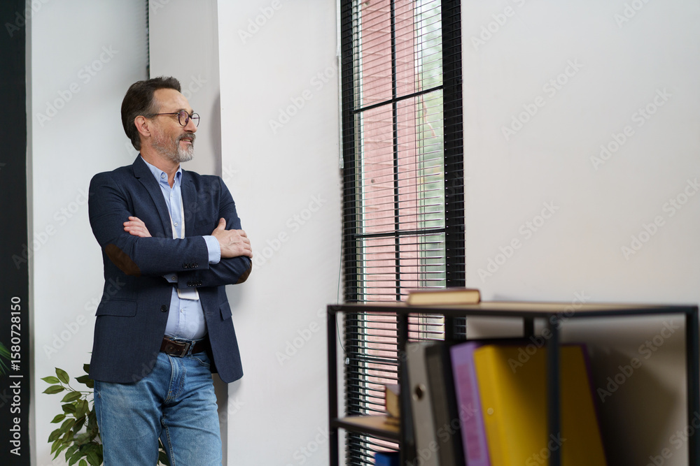 Fototapeta premium A middle-aged man stands confidently with arms crossed by a large window, deep in thought. His stylish attire contrasts with the serene indoor atmosphere, creating a tranquil mood