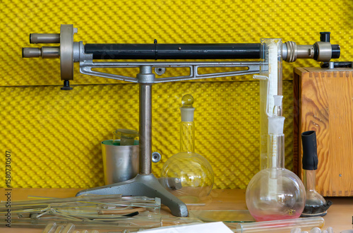 old analytical balance surrounded by glass laboratory vessels and test tubes, placed on a wooden desk against a yellow wall background — scene from a vintage chemistry laboratory.