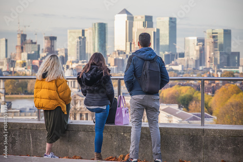 Back view of unidentified tourists appreciating the cityscape of Canary Wharf in London, from Greenwich hill viewing platform 
