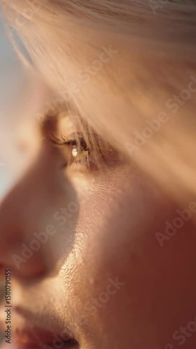 Close-up slow-motion video of a woman with blonde hair blowing in the wind. The sun is shining on her face, and her eyes are closed.
