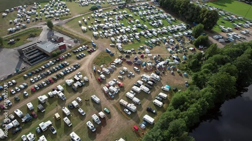 Aerial drone footage flying over a scenic nature campground with multiple RVs, camper vans, and motorhomes parked on a green open space surrounded by trees and natural landscape.

