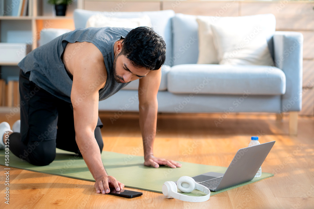 Fototapeta premium Young asian man doing plank exercise in living room at home while watching online workout tutorial on laptop with endurance and motivation, man training plank on yoga mat, digital fitness lifestyle.