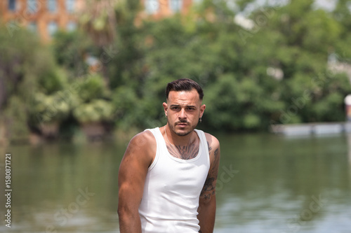 A young, handsome gay man with a beard, tattoos and earrings, wearing a white tank top and black trousers, is leaning on a metal railing by the river. The man is relaxed and resting.