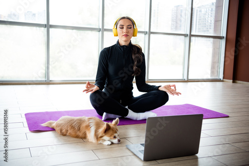 Young woman meditating on a yoga mat with her dog, headphones on, streaming guided meditation session on a laptop