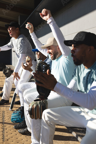 Diverse male baseball teammates cheering while sitting on bench in dugout with bat and gloves