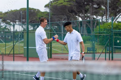 Diverse male tennis teammates exchanging celebratory fist bump at net on green court with racket