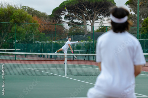 Diverse male tennis opponents wearing headband serving and returning ball on green court by fence