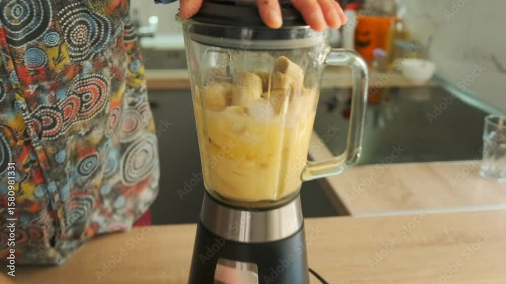 Close-up of a young man in a colorful shirt switching on a countertop blender filled with chopped fruits and water. Blending a healthy homemade smoothie in a cozy modern kitchen. Appliance use.