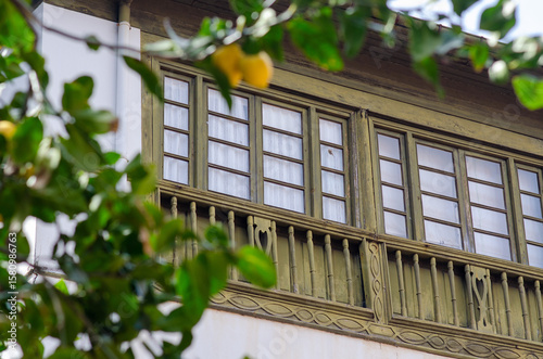 Charming old balcony with intricate wooden details and green frames, partially obscured by leafy lemon branches, capturing rustic Mediterranean architecture and nature.