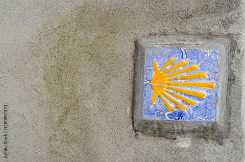 Decorative ceramic tile with yellow scallop shell marker of the Camino de Santiago, embedded in a rustic concrete wall. Symbolizes pilgrimage, travel, and European heritage.