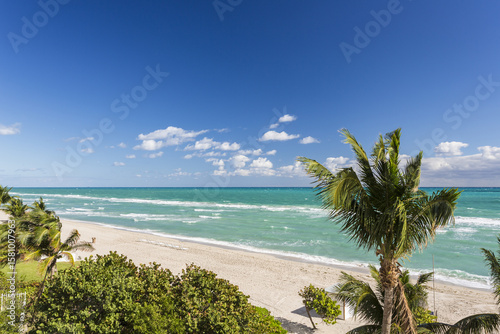 Aerial view of turquoise ocean waves gently kissing the sandy shore under a vast blue sky, punctuated by fluffy white clouds, Miami Beach, Florida, United States.
