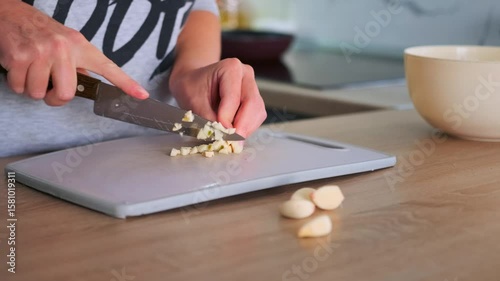 Woman hands chopping raw garlic on a scratched plastic cutting board with a large kitchen knife. Cooking with fresh ingredients at home.
