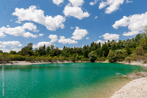 Turquoise lake with pebble shore and clear water, surrounded by forest and blue sky – perfect for recreation, swimming, and relaxing in a peaceful summer nature setting