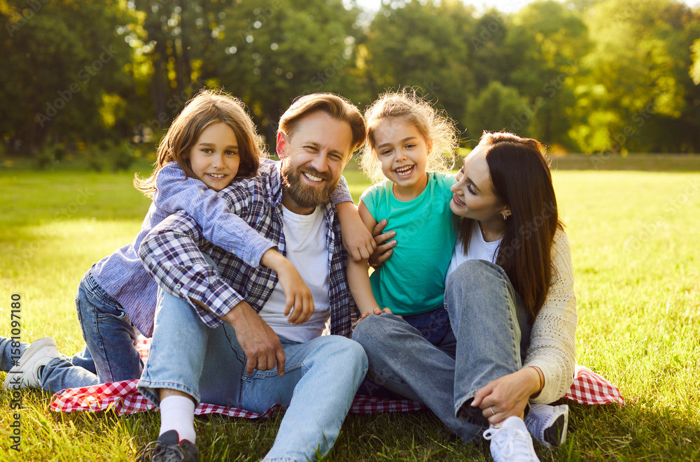 Obraz premium Portrait of happy smiling family with son and daughter sitting on green grass in the summer park and looking cheerful at camera. Mother, father with two kids enjoying sunny day together.
