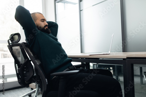 Professional man with beard sitting comfortably in modern office, arms behind his head, taking break from work at desk with open laptop, reflecting relaxed work environment.