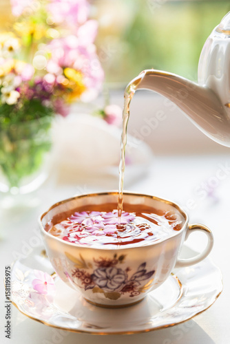 Creative postcard with porcelain cup of tea, teapot and a vibrant bouquet of garden flowers. Elegant tea set in beautiful morning sun light. Good morning and teatime concept. Slow life visual trend.