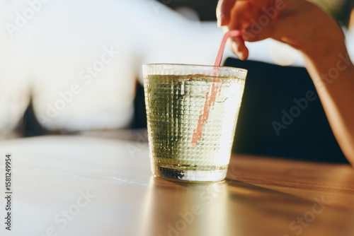 Photos glass of sparkling water with hand stirring pink straw on wooden table in bright
