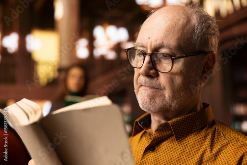 Elderly retired person with spectacles having a reading hobby at the public library, enjoying lecture for leisure activity during retirement. Wrinkled happy man with wisdom and skill, literature. photo